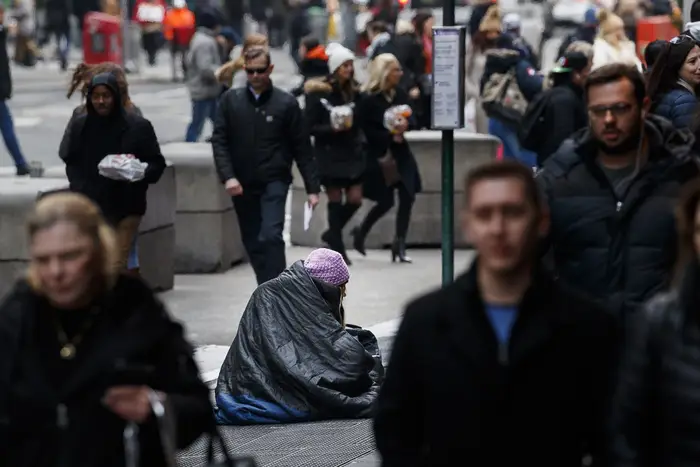 A homeless person sits on a busy NYC street, with many pedestrians passing by, with a sign indicating they are homeless.